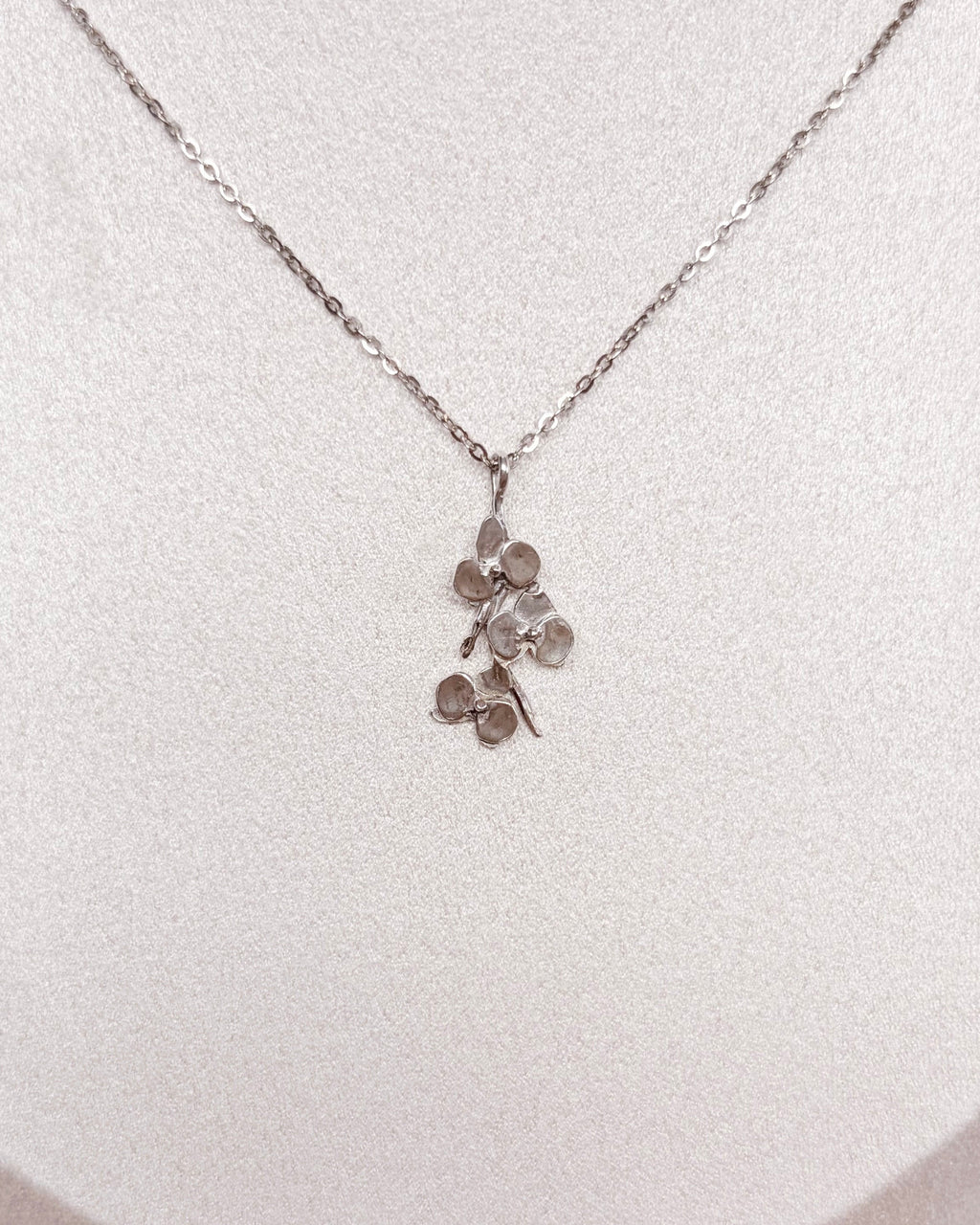 A delicate silver-toned necklace with a small orchid pendant displayed on a white jewelry bust against a warm brown textured background.