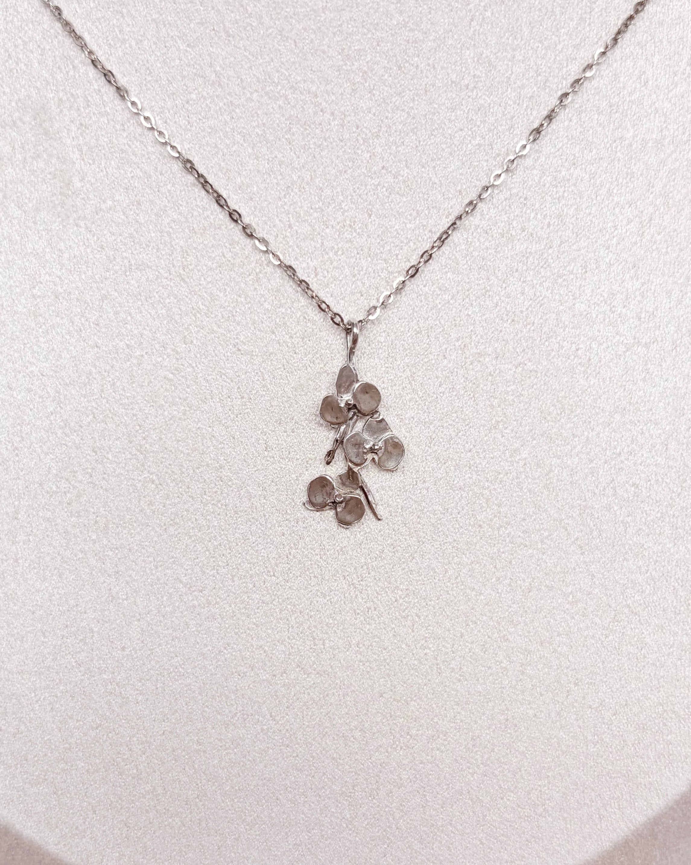 A delicate silver-toned necklace with a small orchid pendant displayed on a white jewelry bust against a warm brown textured background.