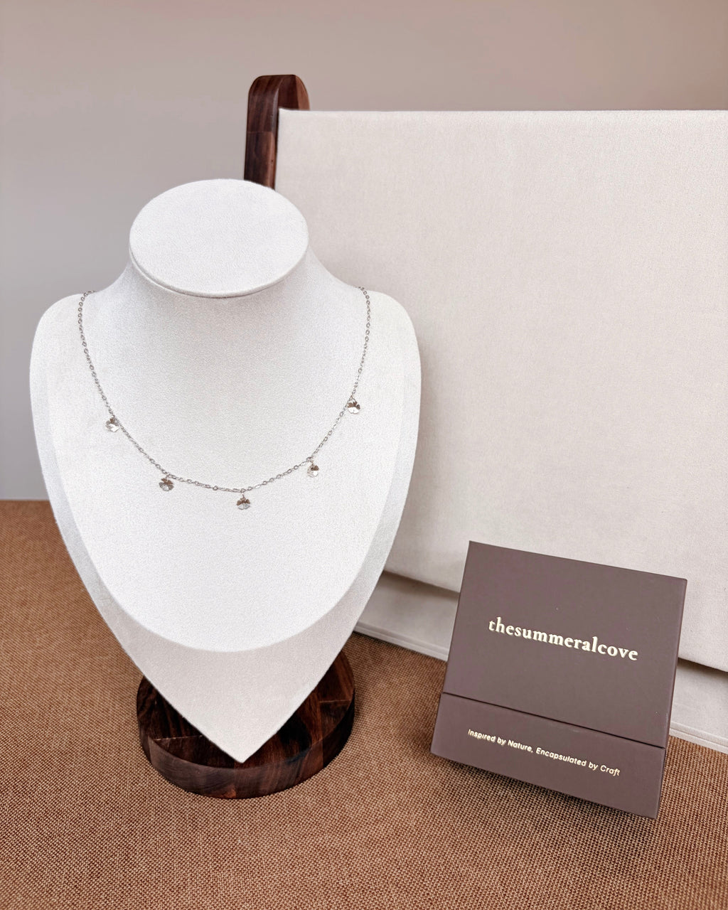 A delicate silver necklace with small dangling charms displayed on a white jewelry bust, with a brown branded box labeled 'thesummercove' beside it on a neutral beige background.