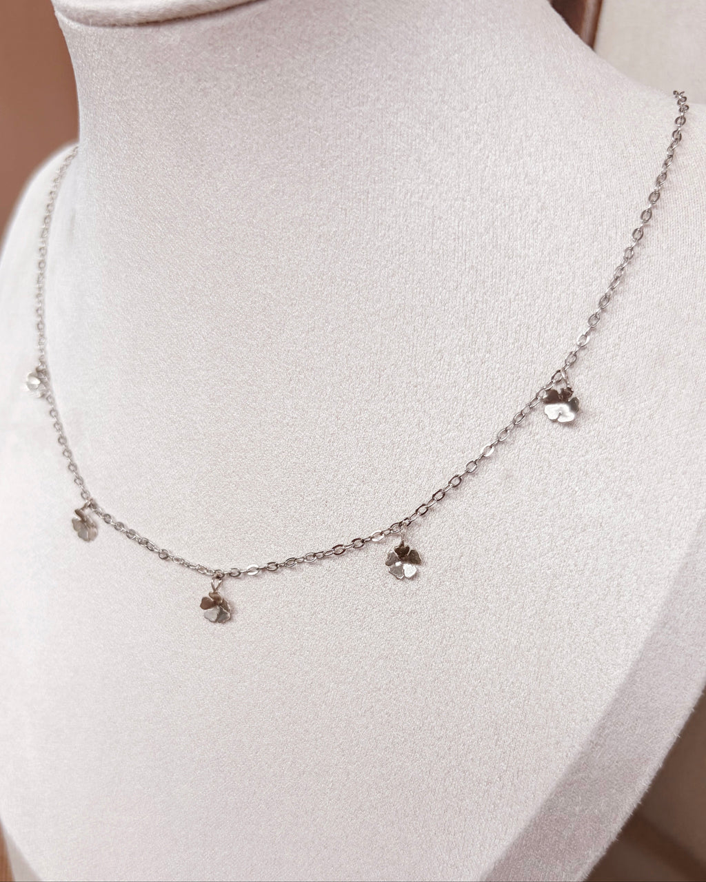 A delicate silver chain necklace with small dangling floral charms, displayed on a white jewelry bust.