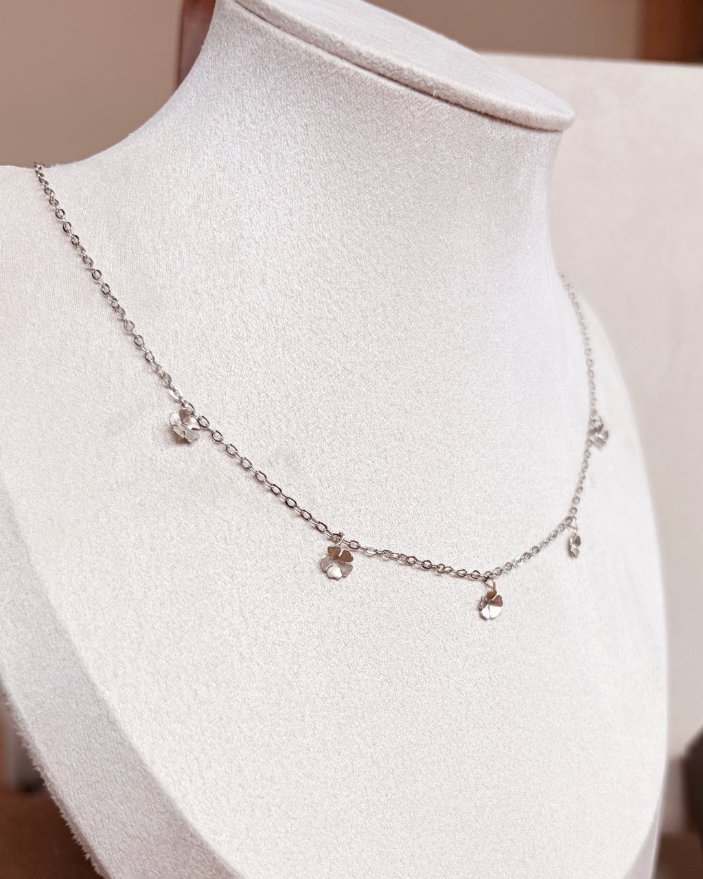 A delicate silver chain necklace with small dangling floral charms displayed on a white jewelry bust. 