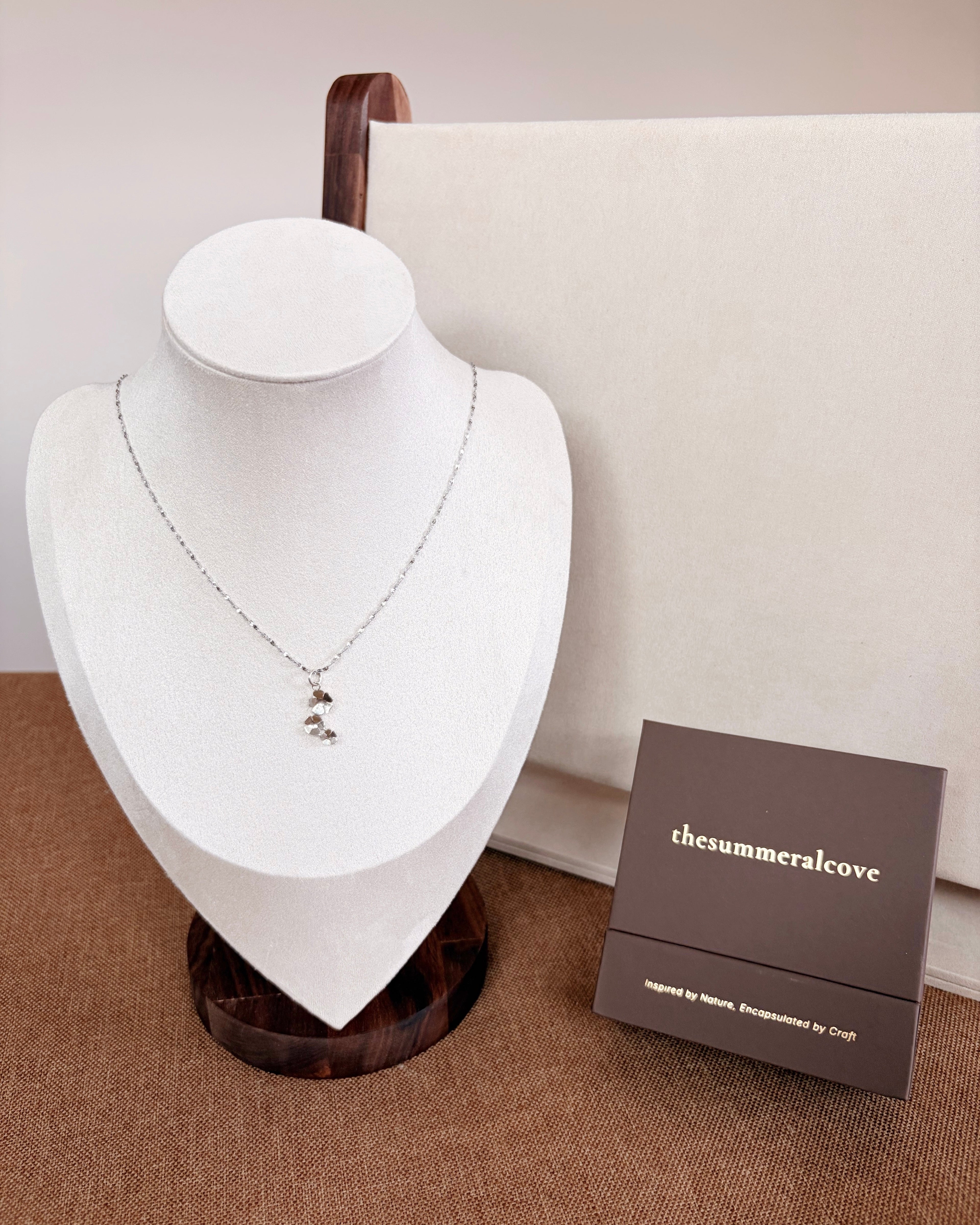 A delicate silver necklace with a small silver flower pendant is displayed on a white jewelry bust. A dark brown brand box labeled 'thesummeralcove' sits next to the bust on a brown textured surface against a neutral beige background.