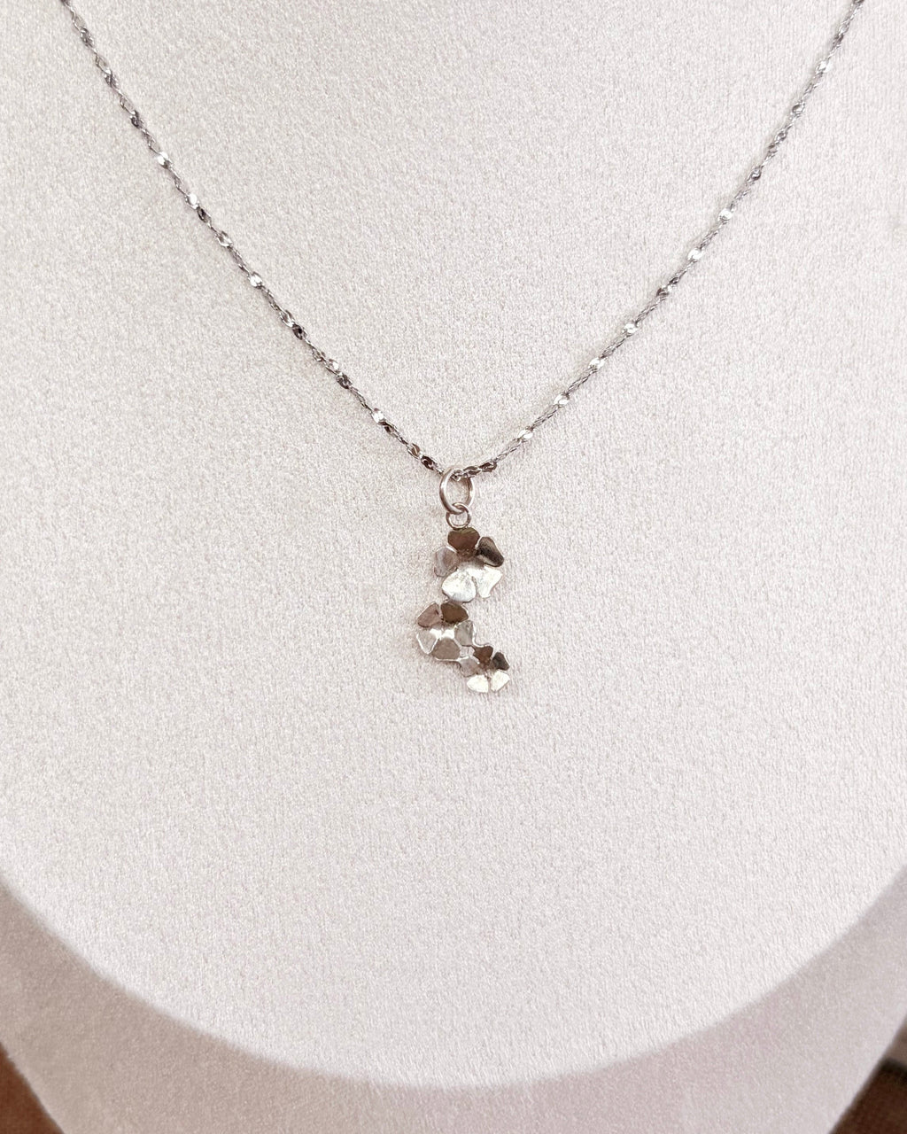 A delicate silver chain necklace with a small silver floral pendant displayed on a white jewelry bust.