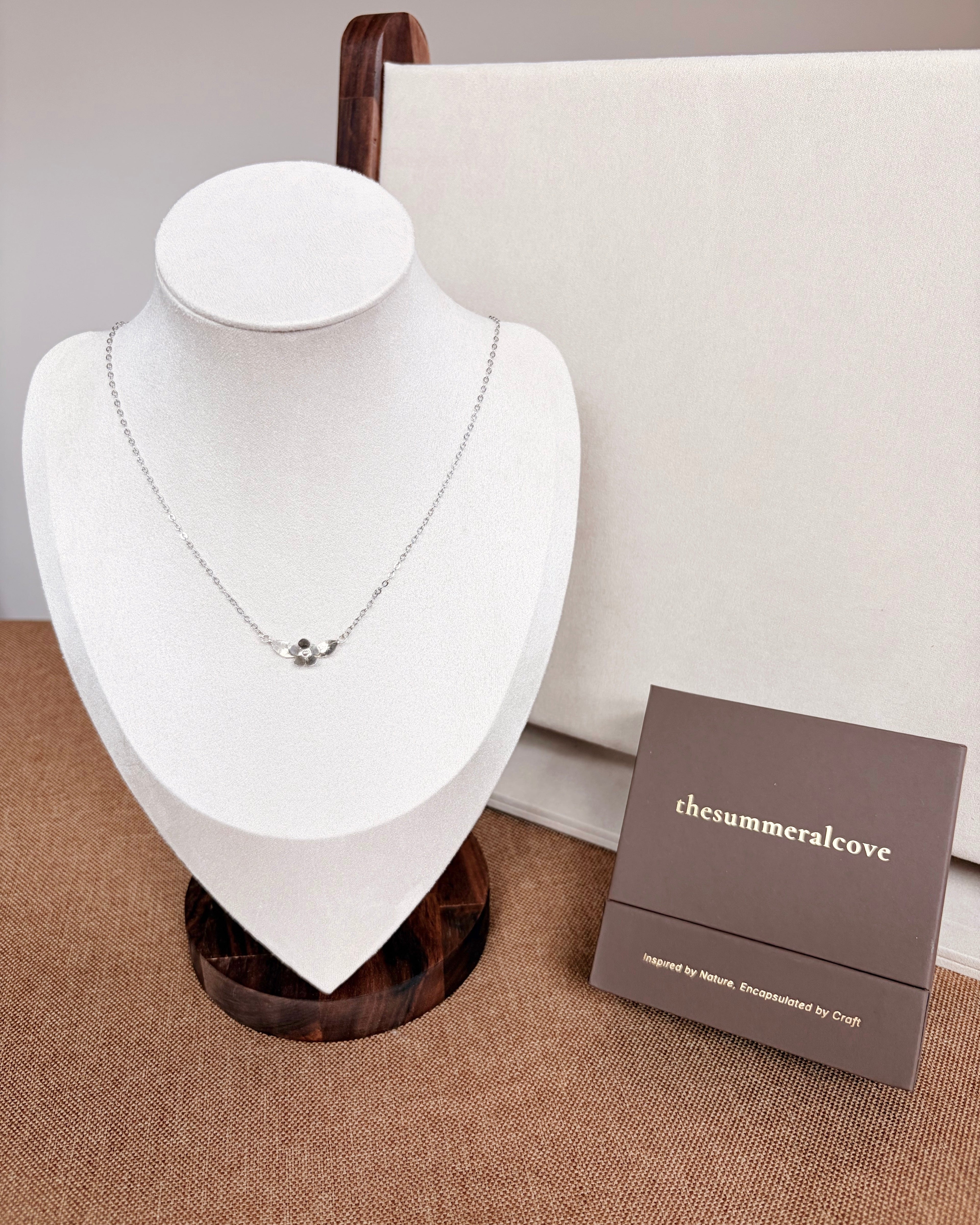 A delicate silver chain necklace with a small plumeria pendant displayed on a white jewelry bust.