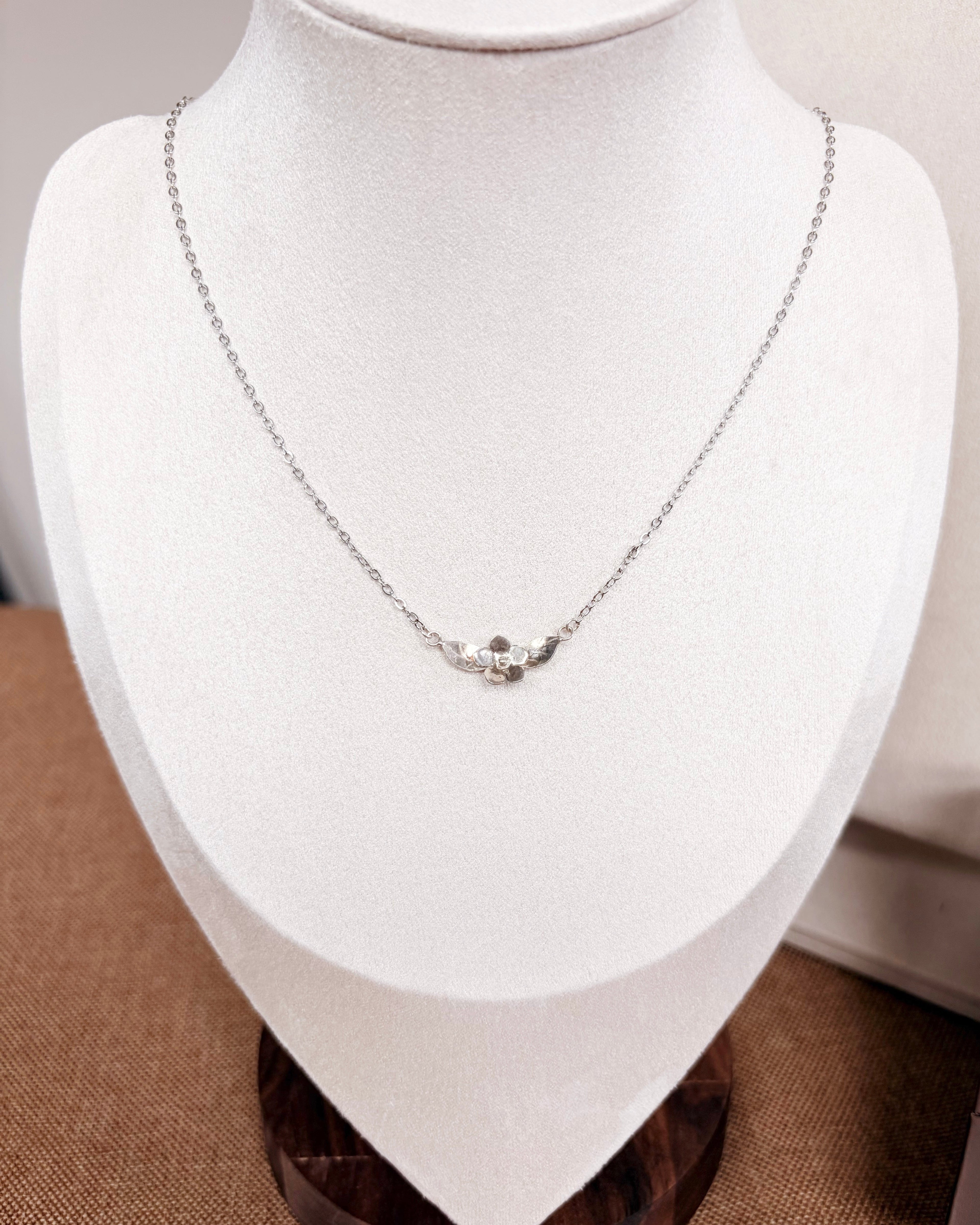 A delicate silver necklace with a small floral plumeria pendant displayed on a white jewelry bust. A brown presentation box with the brand name 'thesummeralcove' sits next to the bust on a textured beige surface.