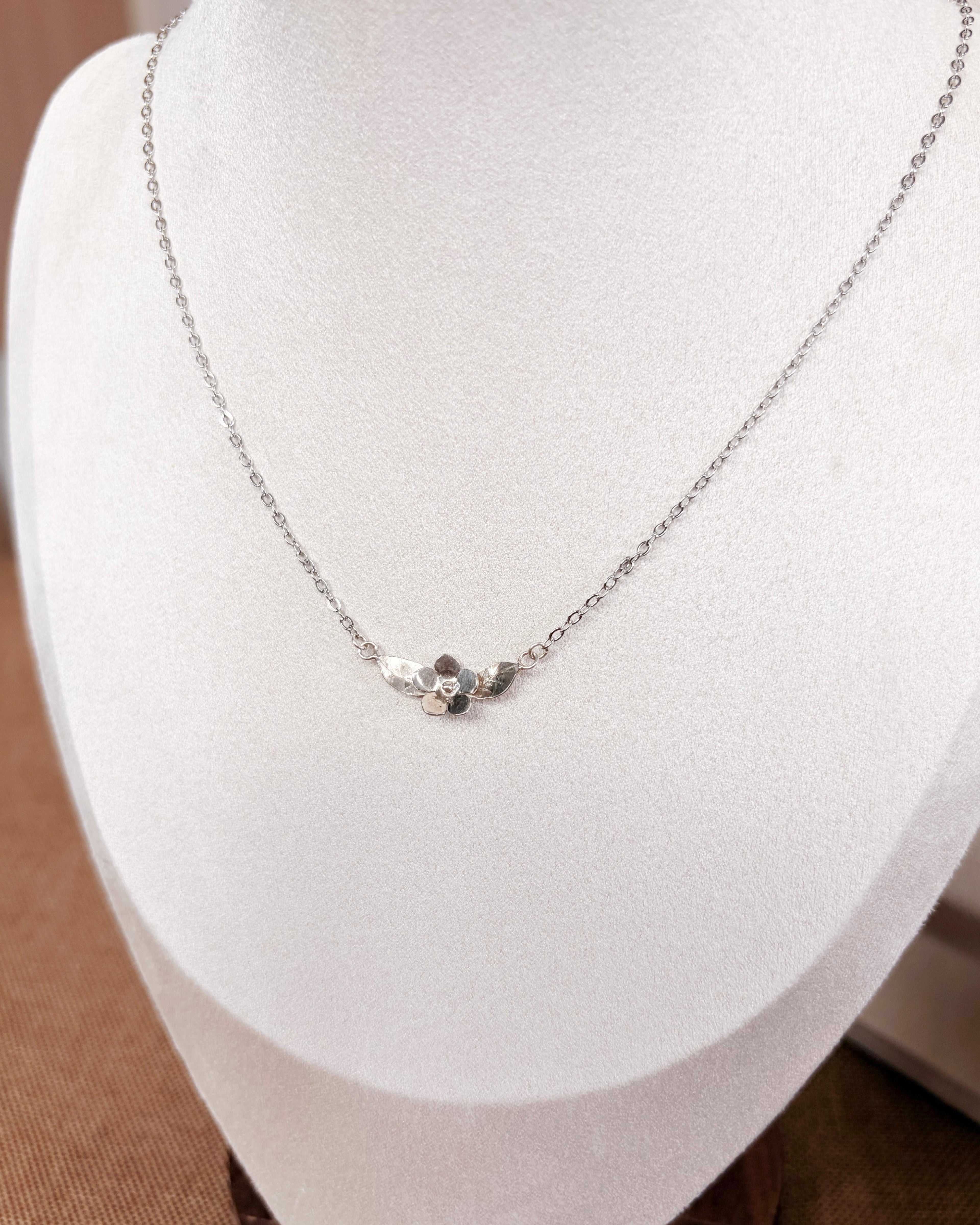 A delicate silver chain necklace with a small silver floral pendant displayed on a white jewelry bust.