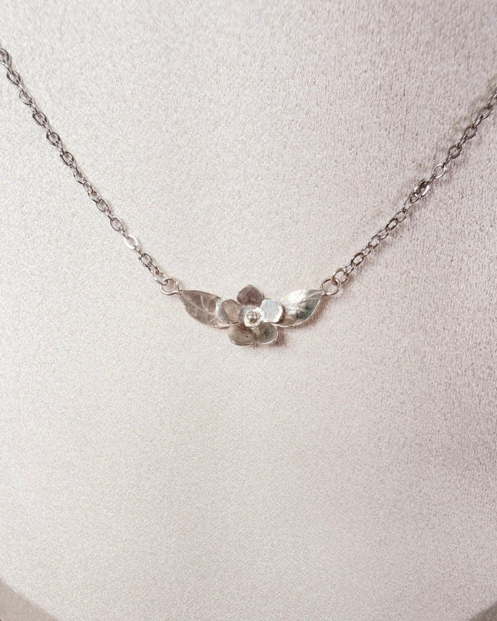 A delicate silver chain necklace with a small textured metal flower pendant, displayed on a textured white jewelry bust.