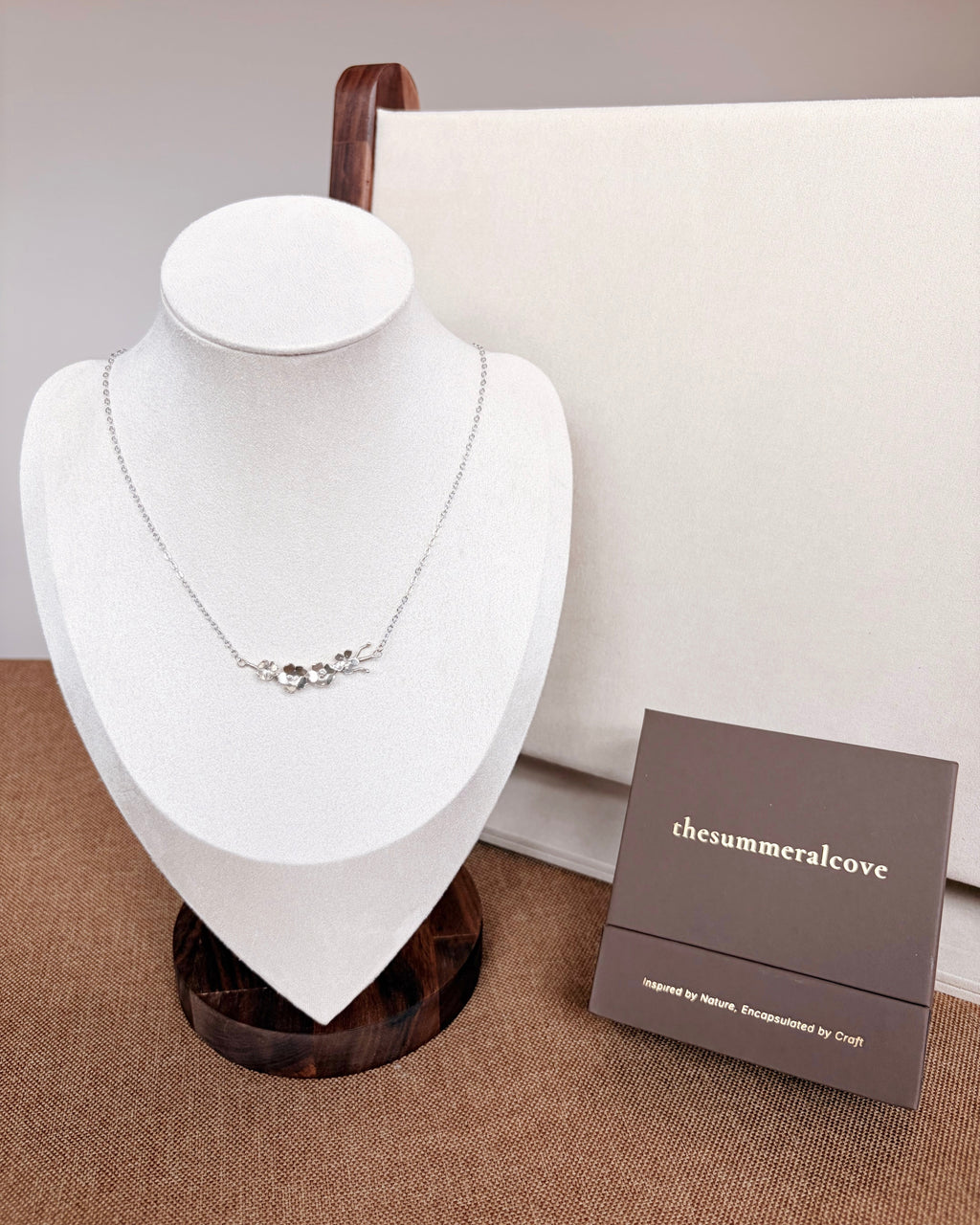 A delicate silver necklace with a small floral and leaf motif displayed on a white jewelry bust. A brown wooden stand and a brown branded gift box labeled 'thesummeralcove' sit beside the bust against a neutral beige background.