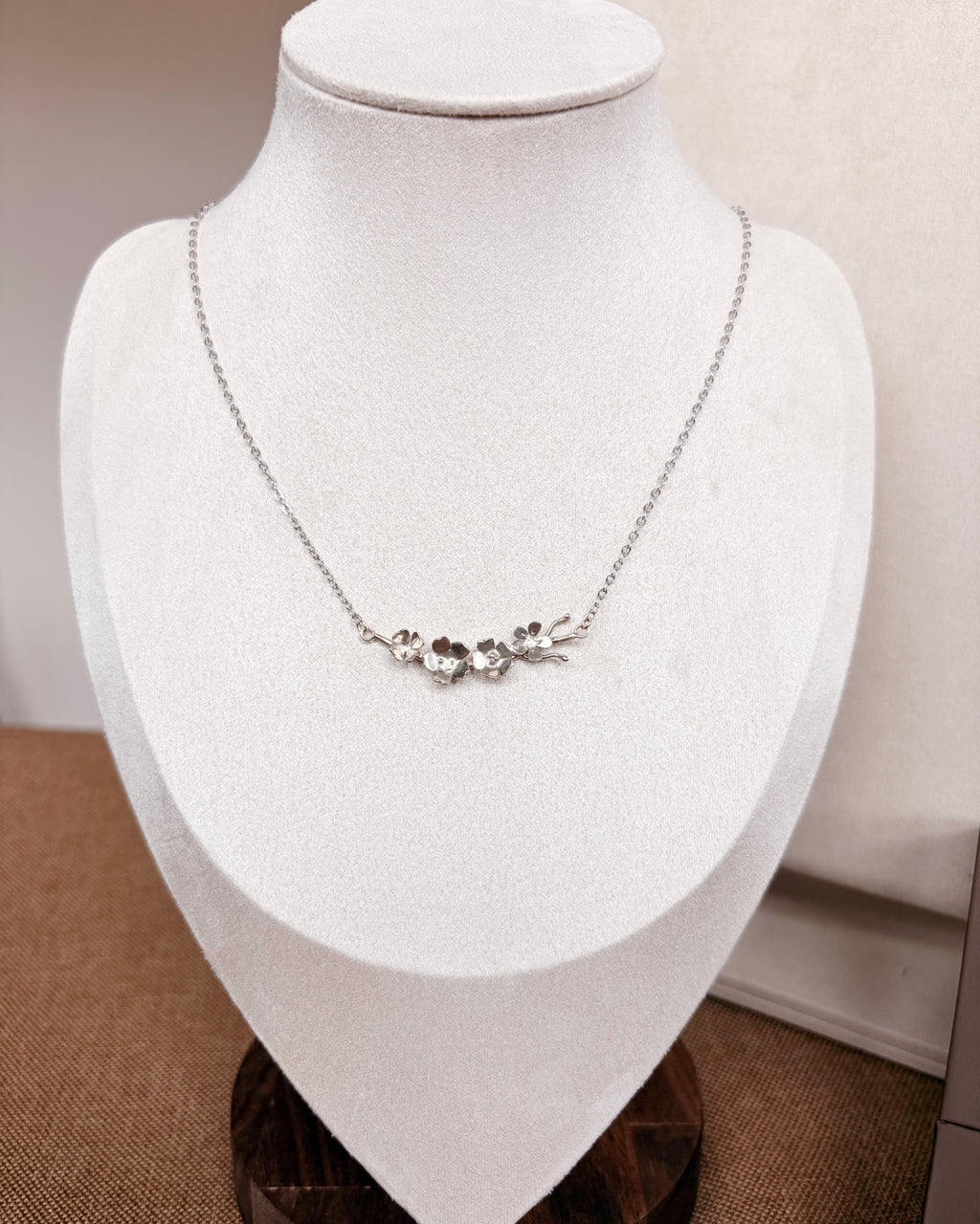 A delicate silver chain necklace with a small floral and branch motif pendant, displayed on a white jewelry bust.