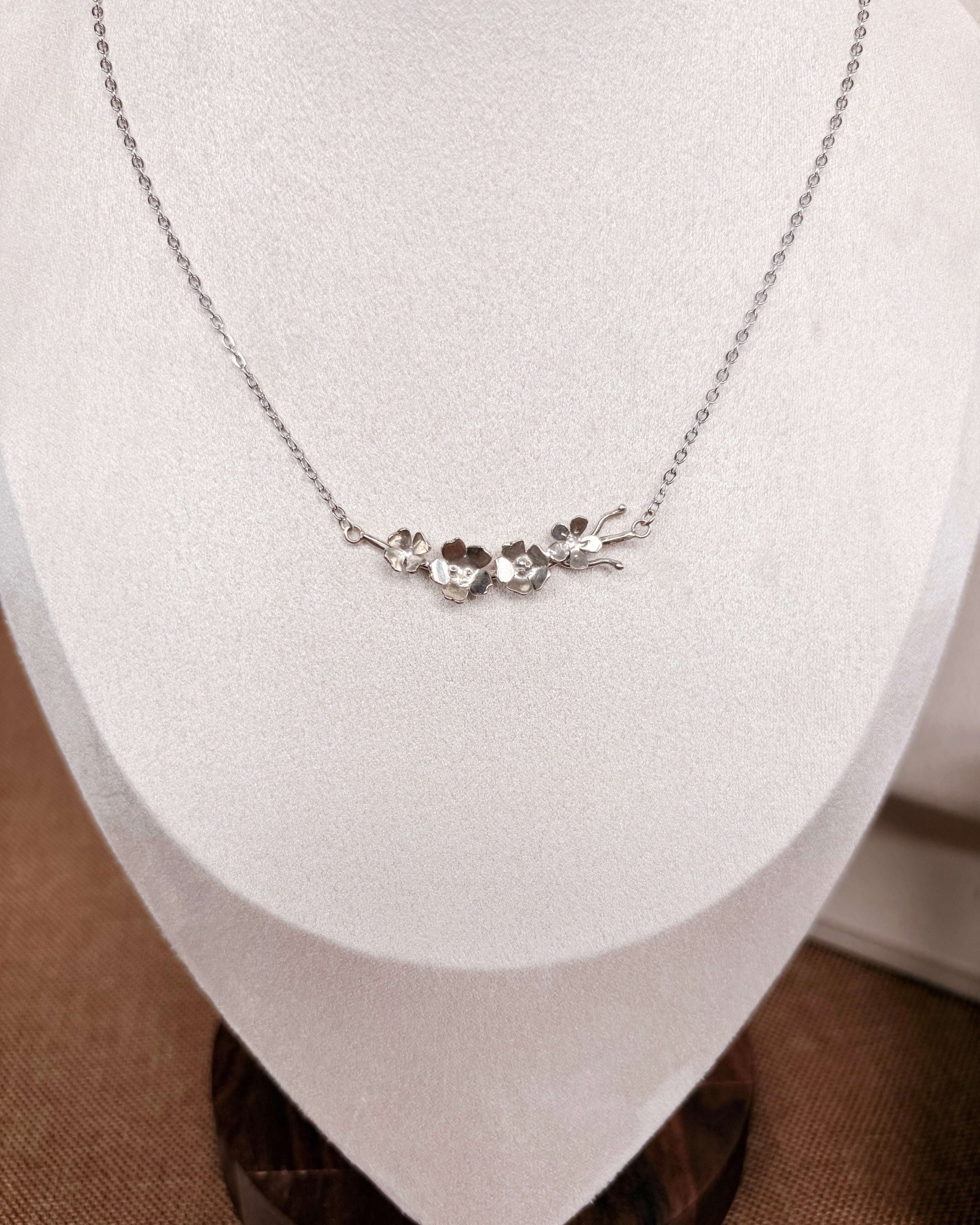 A delicate silver necklace with a small floral and branch pendant, displayed on a white jewelry bust.