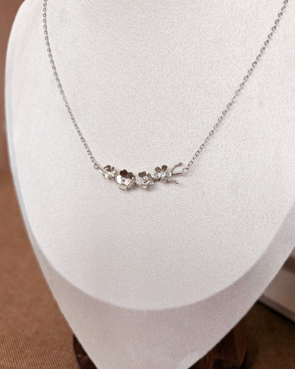 A delicate silver necklace with a small floral and leaf pendant displayed on a white jewelry bust against a warm brown background.