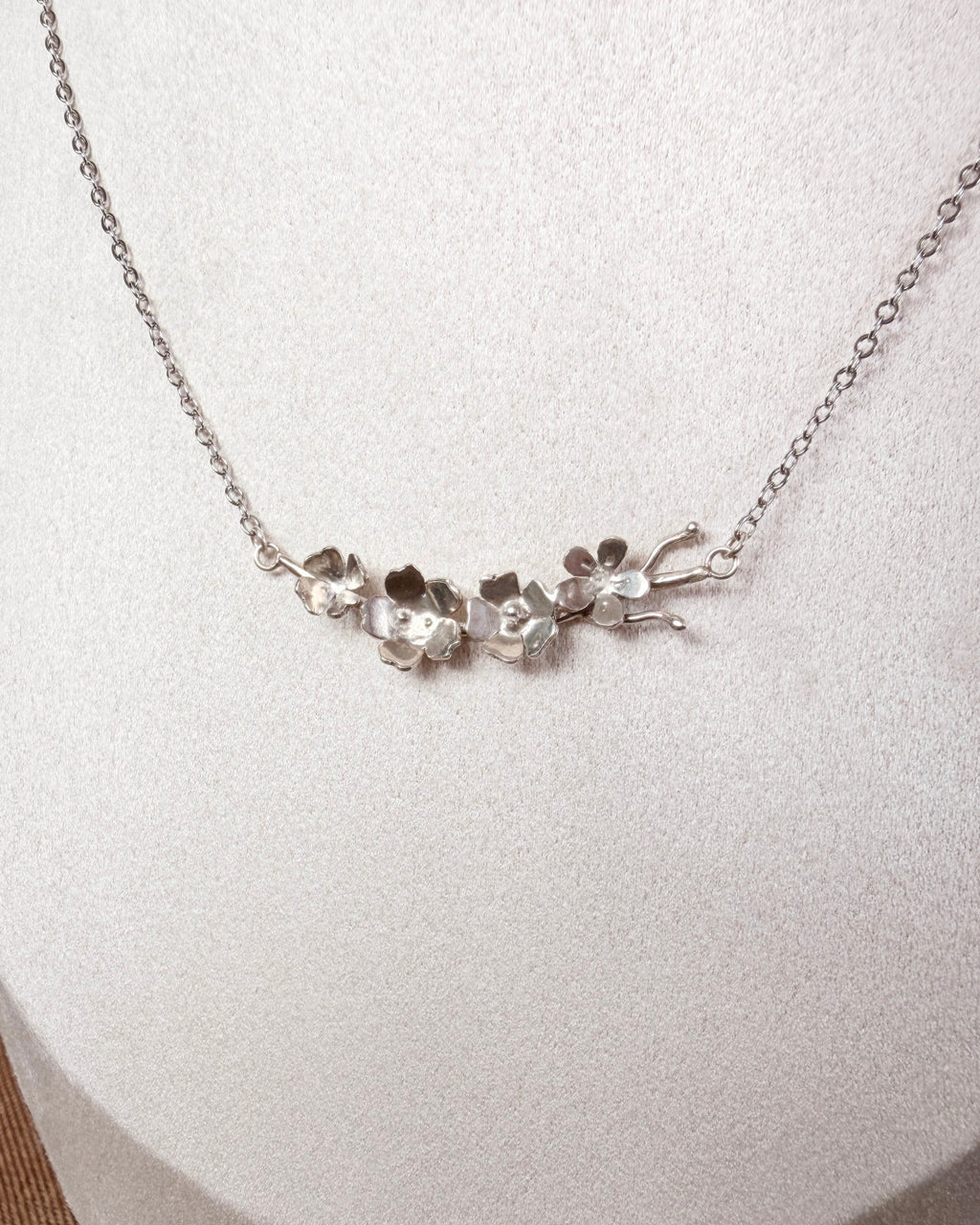 A silver necklace with a delicate chain and a central cluster of small, organic-shaped silver flower charms, displayed on a textured white jewelry bust.