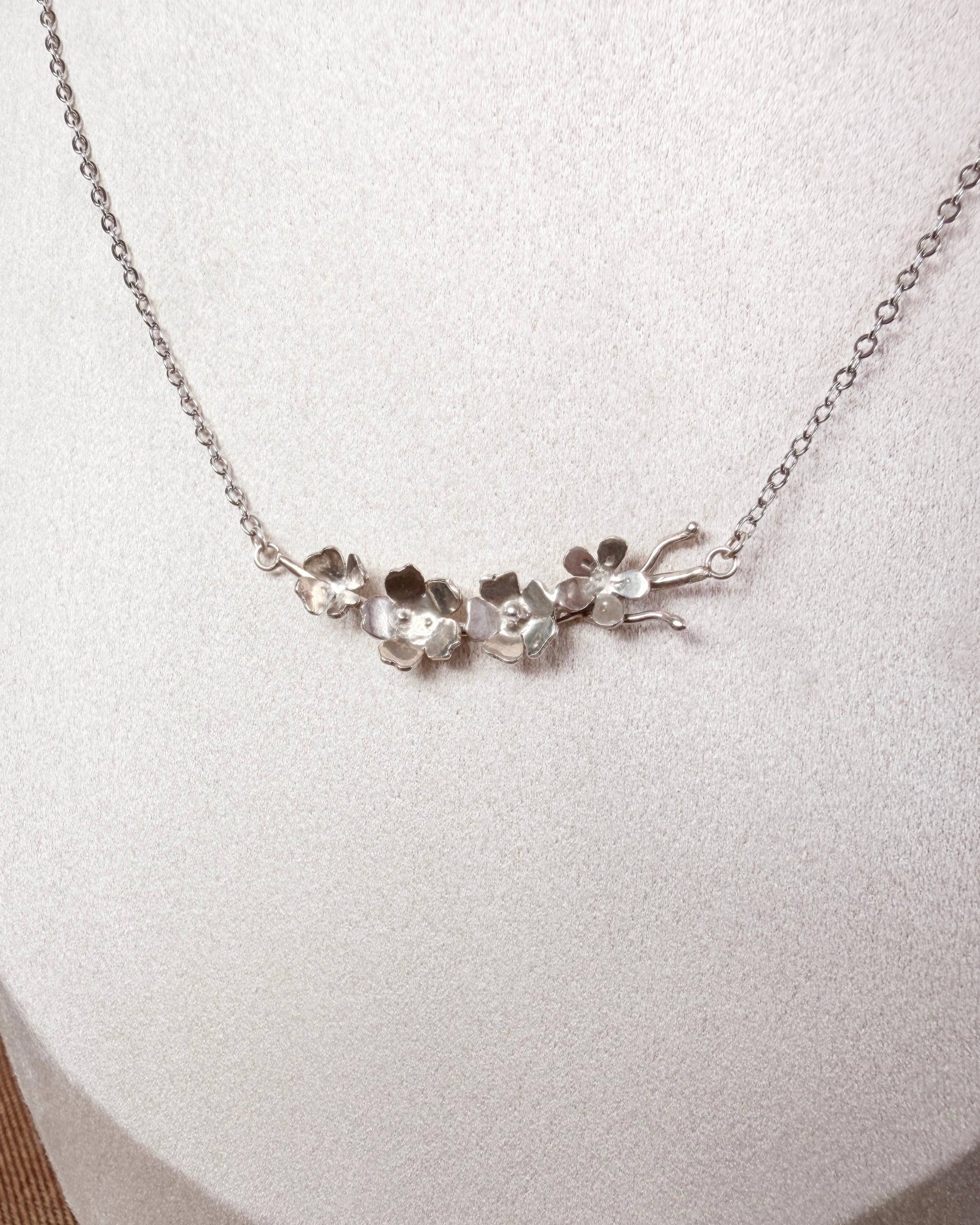 A silver necklace with a delicate chain and a central cluster of small, organic-shaped silver flower charms, displayed on a textured white jewelry bust.