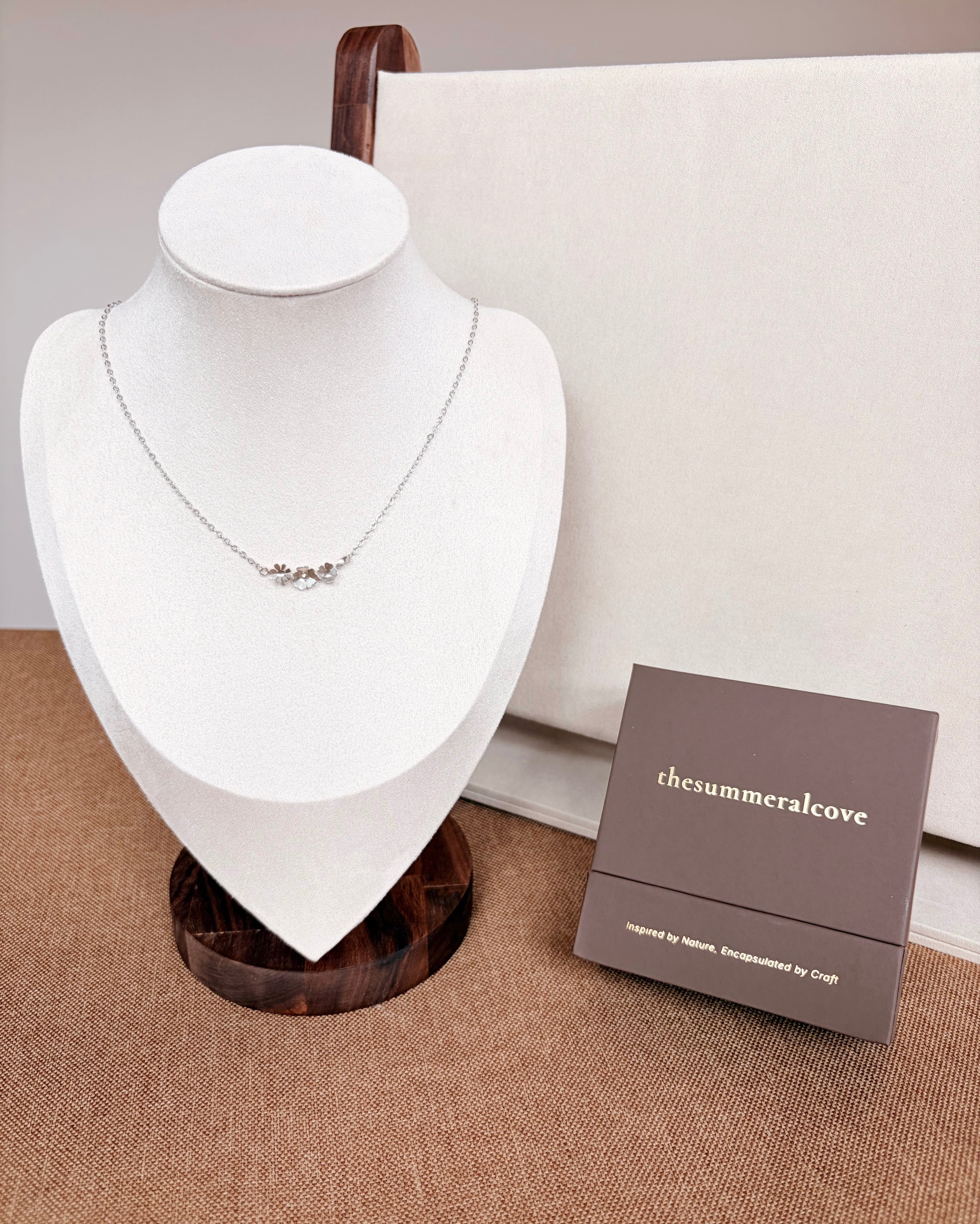 A silver floral necklace displayed on a white jewelry bust, with a small brown jewelry box labeled 'thesummeralcove' beside it on a beige surface.