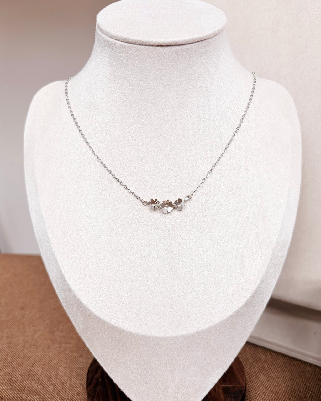 A delicate silver chain necklace with a small central floral or leaf cluster pendant, displayed on a white jewelry bust against a soft beige background.