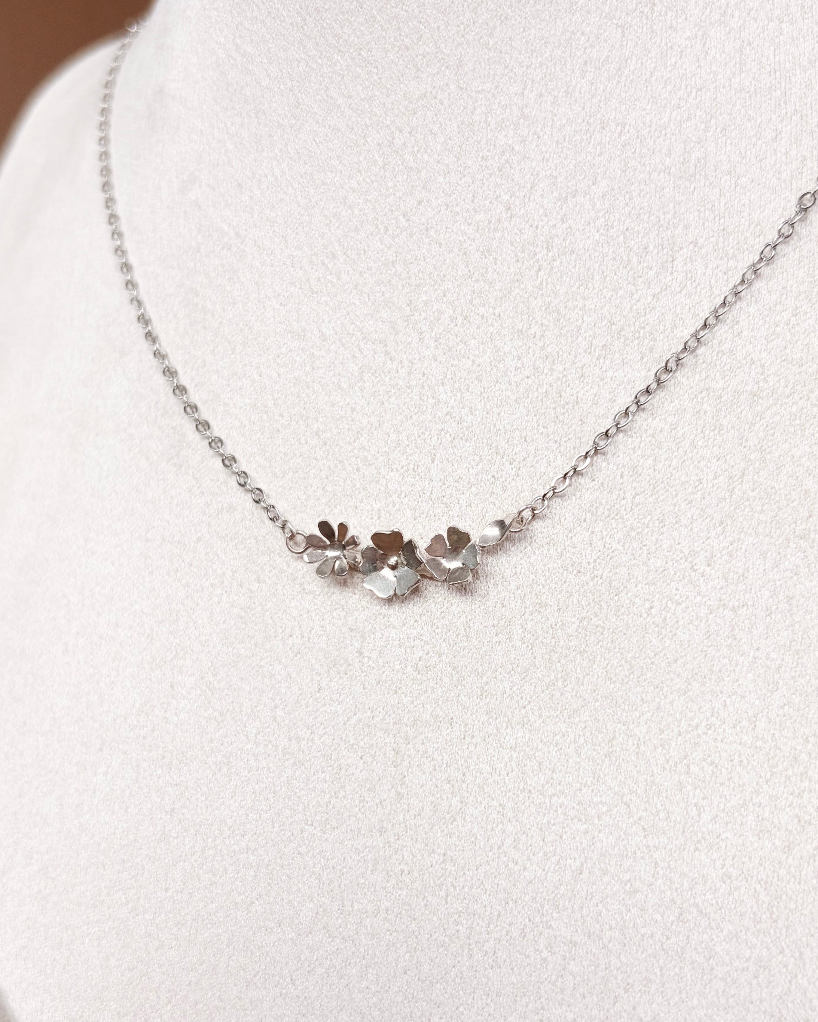 A delicate silver chain necklace with a small central cluster of flowers, displayed on a white jewelry bust.