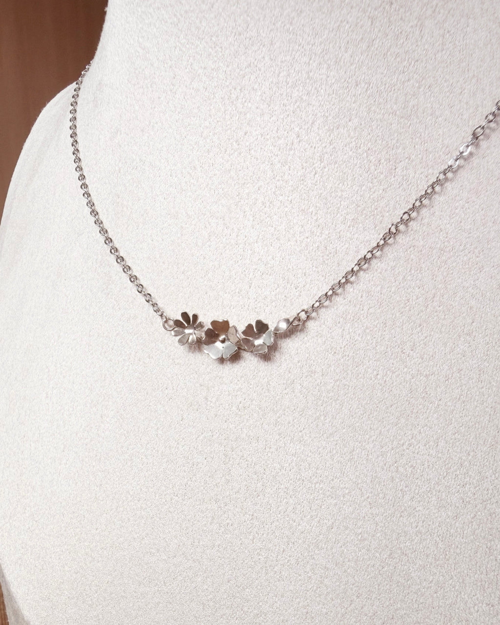 A delicate silver chain necklace with three small flower charms, displayed on a white jewelry bust.