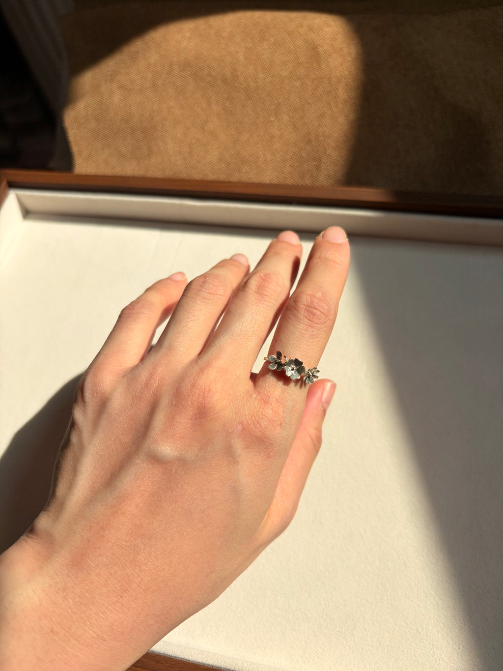 A hand wearing a silver-toned ring with small dainty wildflowers, resting on a cream-colored jewelry box lid under warm natural sunlight.