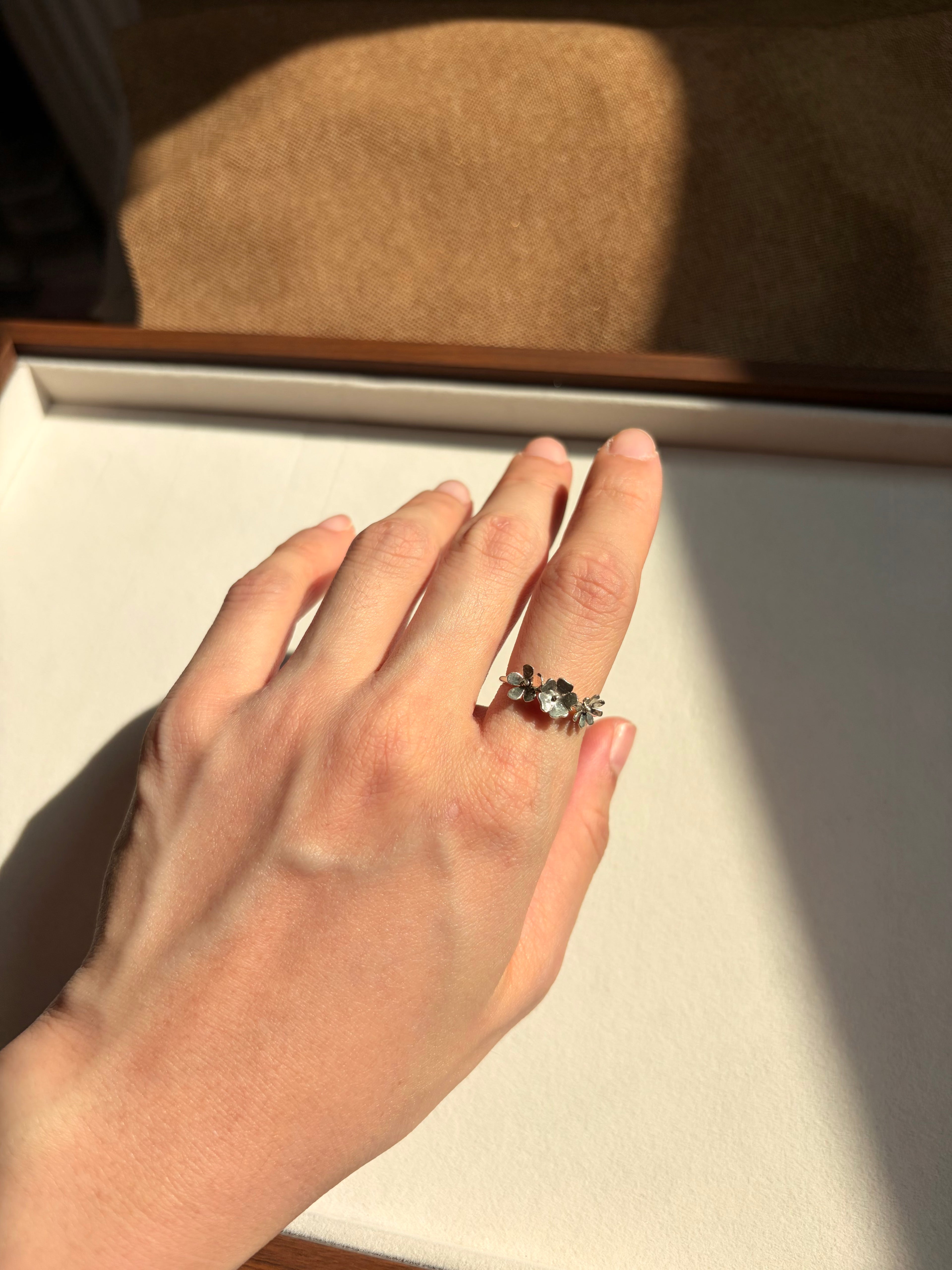 A hand wearing a silver-toned ring with small dainty wildflowers, resting on a cream-colored jewelry box lid under warm natural sunlight.