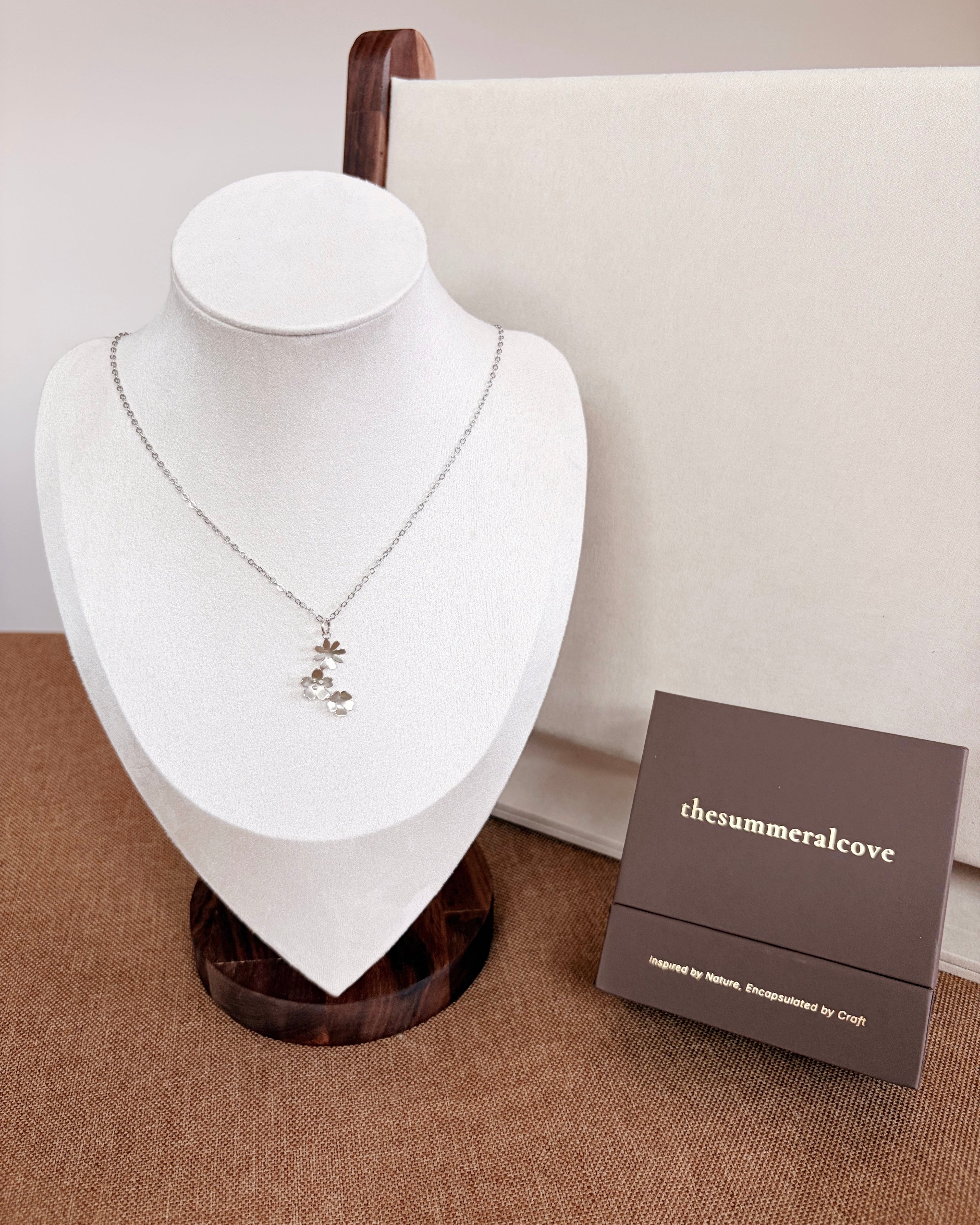 A silver-toned necklace with a small floral or leaf pendant displayed on a white jewelry bust. A dark brown branded box labeled 'thesummeralcove' sits beside the bust on a textured tan surface, with a cream backdrop and wooden stand behind.