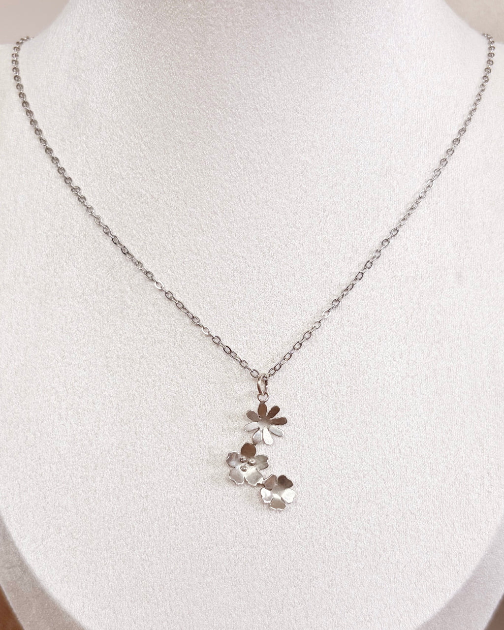 A delicate silver chain necklace with a small cluster of wildflowers pendants displayed on a white fabric jewelry bust.