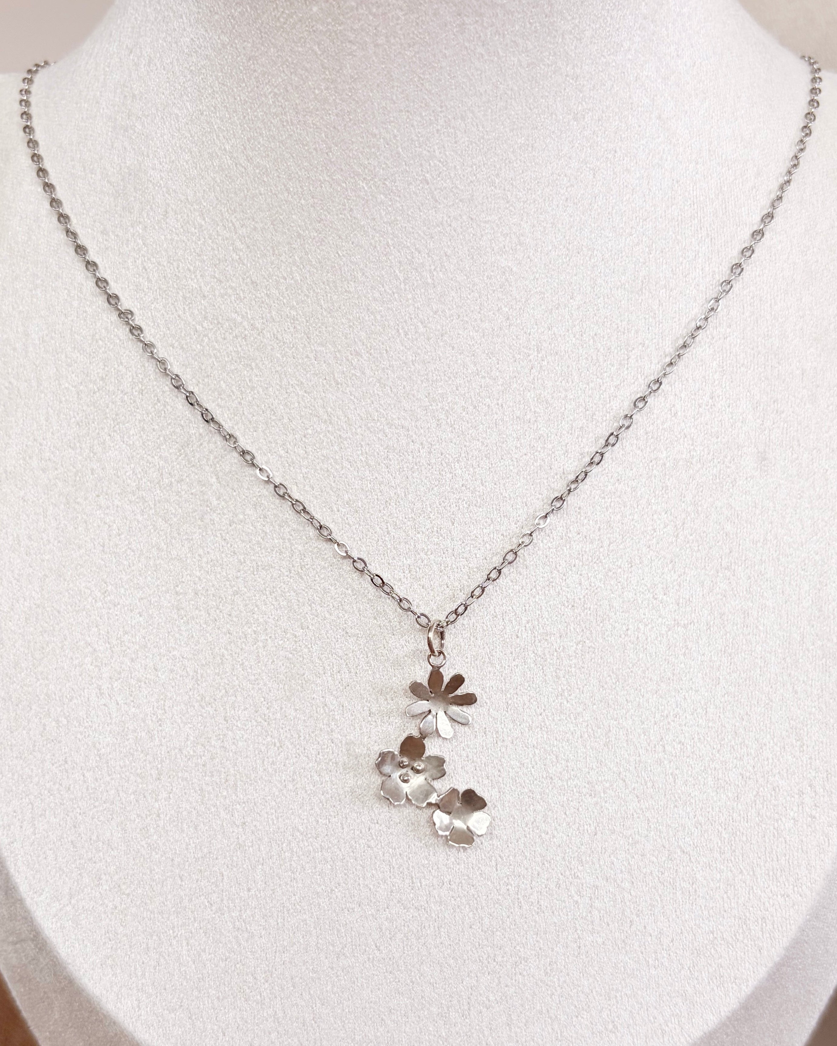 A delicate silver chain necklace with a small cluster of wildflowers pendants displayed on a white fabric jewelry bust.