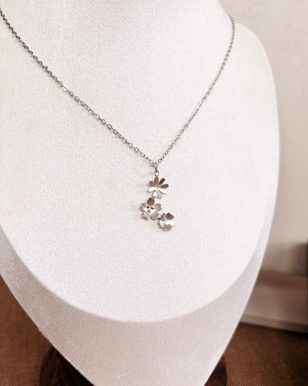 A silver floral pendant necklace displayed on a white jewelry bust against a soft beige background.